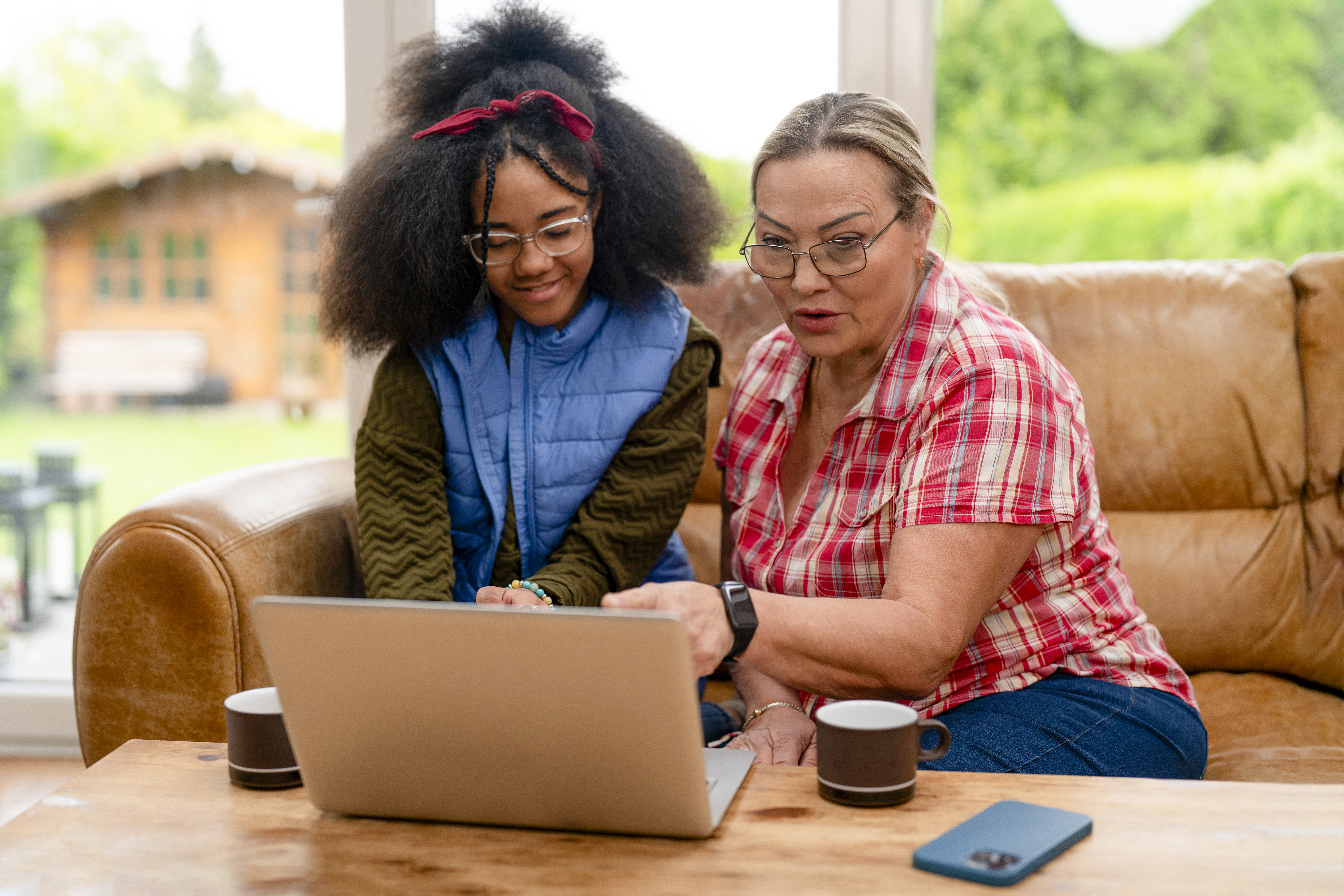 A young girl and an older woman are sitting on a sofa in a living room. They are using a laptop together and sharing ideas. Coffee mugs are on the table.